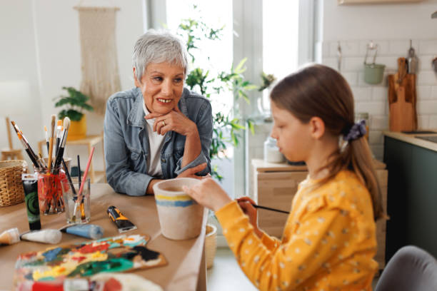 A senior woman resident and her granddaughter smiling in a cozy, light-filled studio painting a pot